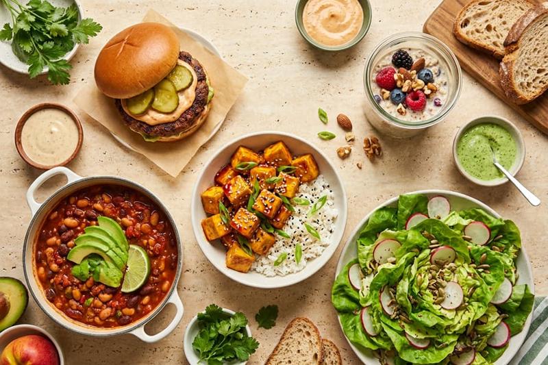 A spread of plant-based dishes: a burger, sticky tofu over rice, bean chili, a big salad and berry oats